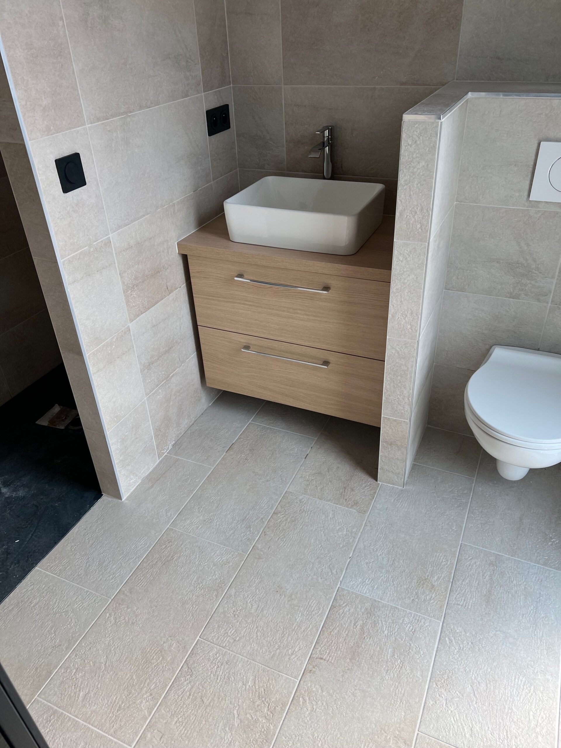 Un plombier répare une canalisation derrière des toilettes dans une salle de bain carrelée de gris.